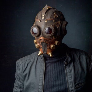 May include: A steampunk-style mask made of brown and tan leather, featuring large, round goggles with red lenses. The mask has brass accents and a detailed, industrial design, with a dark background.