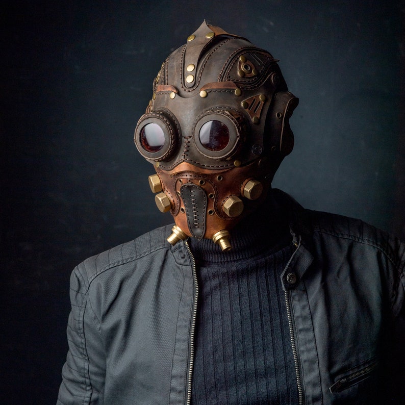 May include: A steampunk-style mask made of brown and tan leather, featuring large, round goggles with red lenses. The mask has brass accents and a detailed, industrial design, with a dark background.