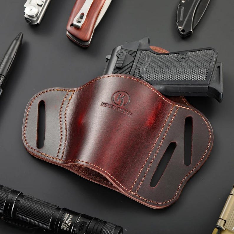May include: A burgundy leather holster with a stitched border, holding a black handgun. The holster has cutouts and the brand name "KOSTPATE" is visible. Other tools are in the background.