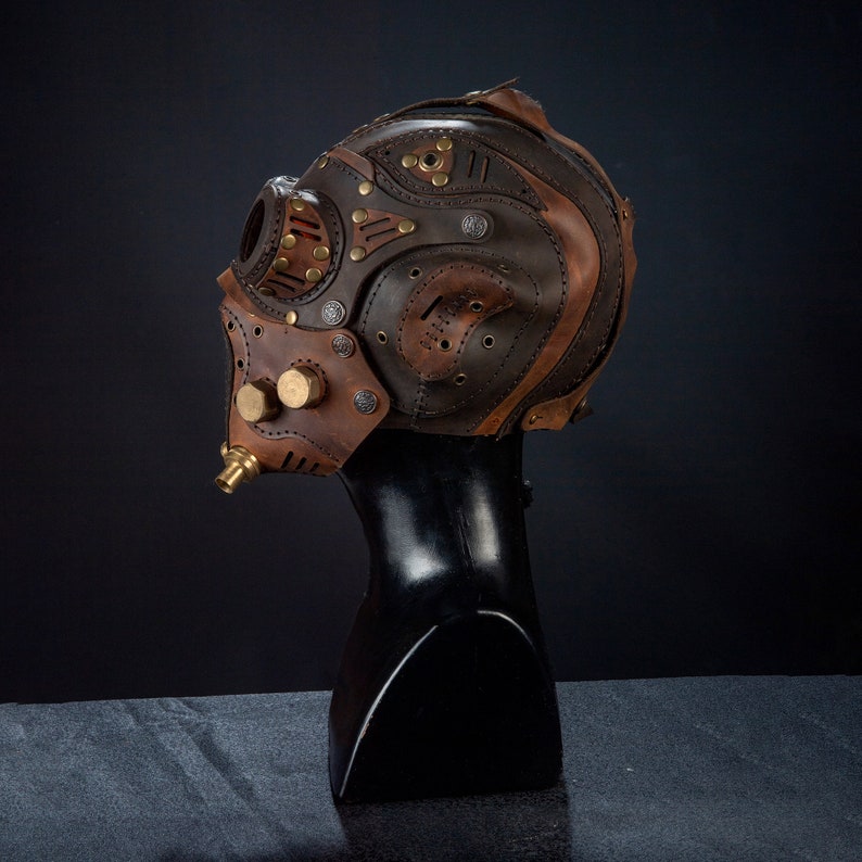 May include: A steampunk-style leather mask in shades of brown, tan, and copper. The mask features rivets, circular accents, and a brass nozzle. It is displayed on a black mannequin head against a dark background.