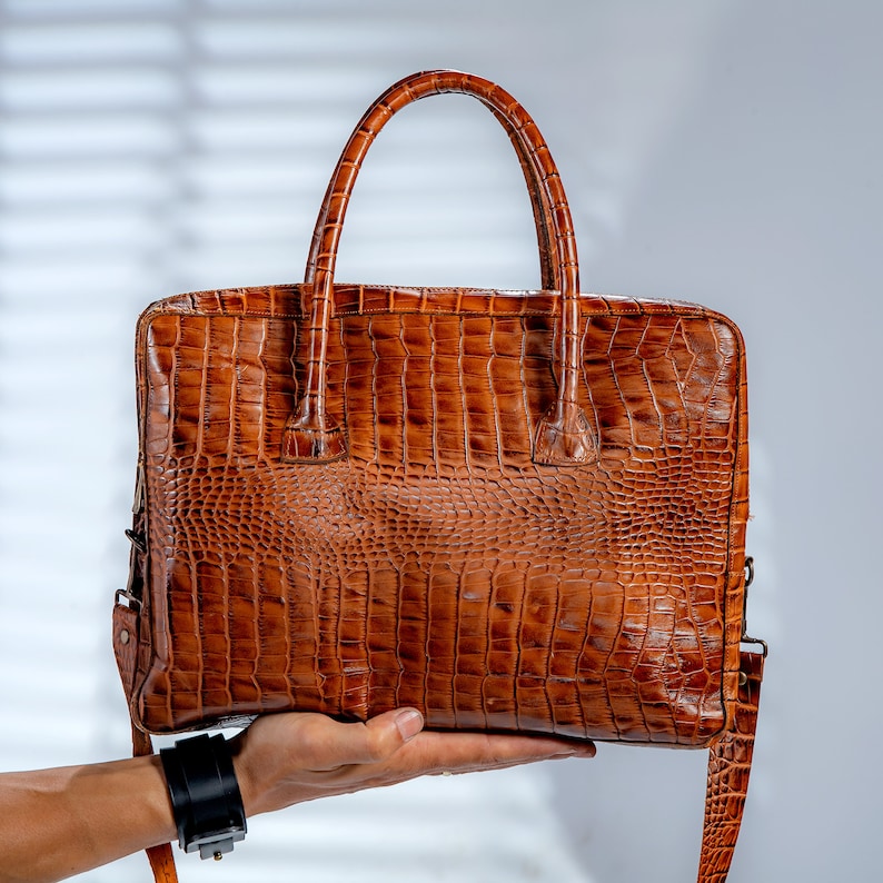 May include: A brown leather briefcase with a crocodile skin texture. The bag has a top handle and a detachable shoulder strap. The briefcase is held up by a person's hand.