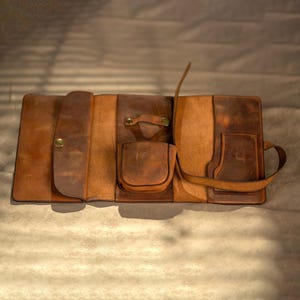 May include: Open, brown leather organizer with multiple pockets and compartments. Features a snap closure, a small handle, and a long strap. The leather has a distressed look with variations in color.