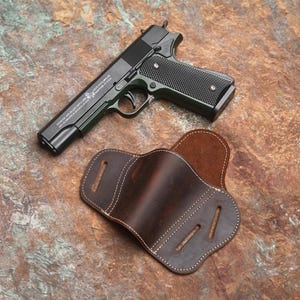 May include: A black handgun with a textured grip and a brown leather holster. The holster has white stitching and a curved design. The gun is lying next to the holster on a brown surface.