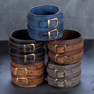 May include: A stack of leather wrist cuffs in various colors: blue, black, brown, and gray. Each cuff features two straps with brass-colored buckles. The cuffs are stacked on a dark surface, showcasing their design and texture.