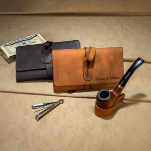 May include: Two leather tobacco pouches, one brown and one tan, with a clasp closure. A wooden pipe with a black stem and a silver pipe tool are also present. The tan pouch has the name "Kimmie Jo Thacker" engraved on it.