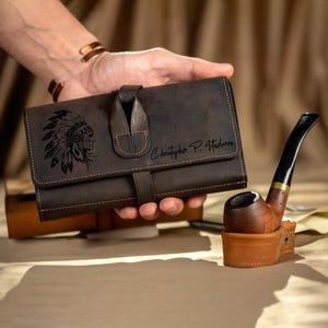 May include: A dark brown leather tobacco pouch with a flap closure, featuring a Native American chief design and the name "Christopher P. Attadonna." A wooden pipe with a black stem rests nearby.