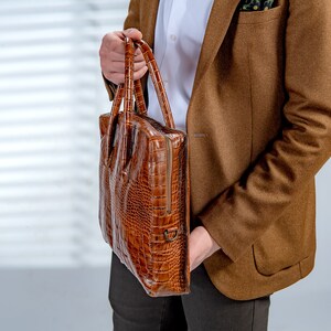 May include: A brown leather briefcase with a crocodile skin texture. The briefcase has a zip closure and two handles. The person is wearing a brown blazer and a white shirt.