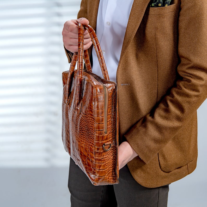 May include: A brown leather briefcase with a crocodile skin texture. The briefcase has a zip closure and two handles. The person is wearing a brown blazer and a white shirt.