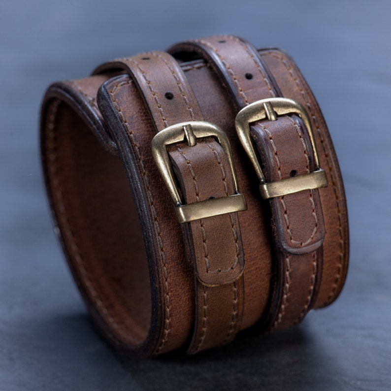 May include: A brown leather cuff bracelet with two straps and brass buckles. The wide bracelet is made of distressed leather and has a stitched design. The cuff is a stylish accessory.