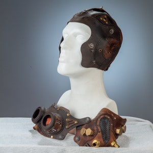 May include: A brown leather aviator helmet and goggles set. The helmet features stitched panels, rivets, and a top flap. The goggles have circular lenses, brass accents, and a steampunk aesthetic. The set is displayed on a white mannequin head.