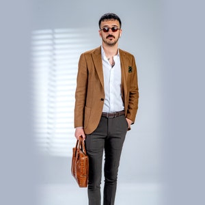 May include: A man in a brown blazer, white shirt, and grey trousers, holding a brown leather briefcase. The blazer has a two-button closure. The outfit is completed with a brown belt and sunglasses. The briefcase has a crocodile skin pattern.