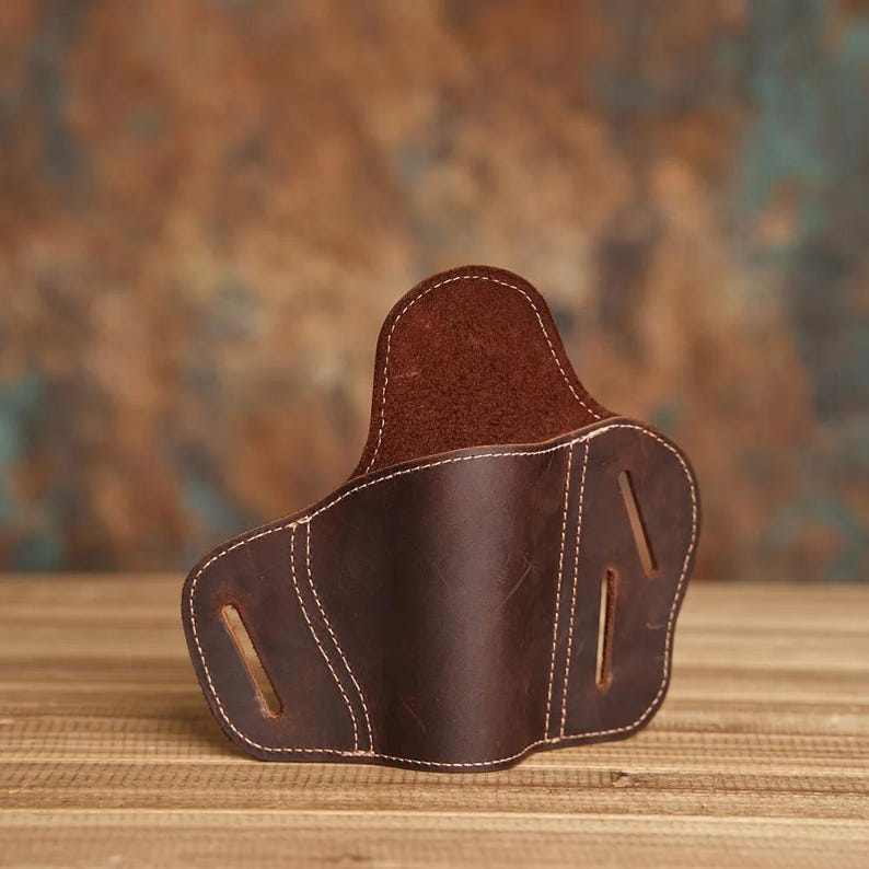 May include: A brown leather holster with white stitching. The holster has a curved shape with three slots on the side. The leather has a distressed appearance, and the background is a blurred mix of brown and blue.