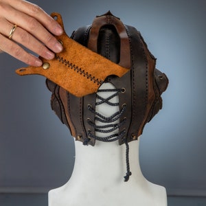 May include: A handcrafted leather mask in shades of brown, featuring a lace-up front and a unique, textured design. The mask includes a separate, tan leather piece with decorative stitching and brass rivets. The mask is displayed on a white mannequin head.