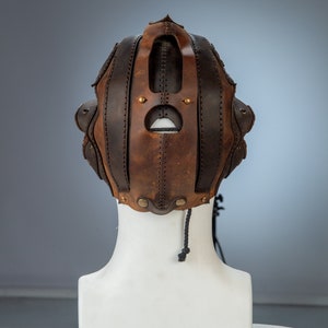 May include: A brown and black leather mask with a central eye opening and riveted accents. The mask has a series of vertical and horizontal leather straps, and a black cord hangs from the bottom. The mask is displayed on a white mannequin head.
