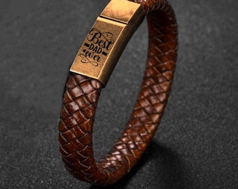 May include: Brown leather braided bracelet with a rectangular gold-colored clasp. The clasp is engraved with the words "Best Dad Ever" in a decorative font. The bracelet is a dark brown color.