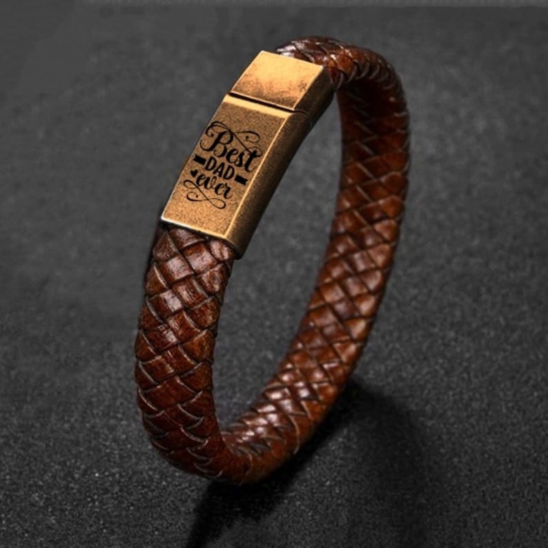 May include: Brown leather braided bracelet with a rectangular gold-colored clasp. The clasp is engraved with the words "Best Dad Ever" in a decorative font. The bracelet is a dark brown color.