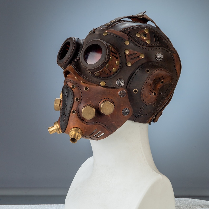 May include: A brown and tan leather steampunk mask with brass accents and goggles. The mask features intricate stitching, rivets, and a unique design. The goggles have red lenses. The mask is displayed on a white mannequin head.