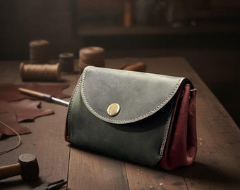 May include: A handcrafted leather pouch in shades of gray and burgundy, secured with a brass button. The pouch is on a wooden surface, with leatherworking tools in the background, suggesting a handmade product.
