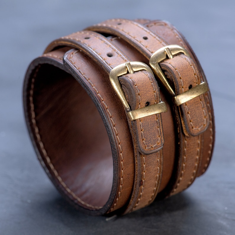 May include: Brown leather cuff bracelet with two straps and brass buckles. The cuff is approximately 2 inches wide and features detailed stitching. The bracelet is designed to wrap around the wrist.