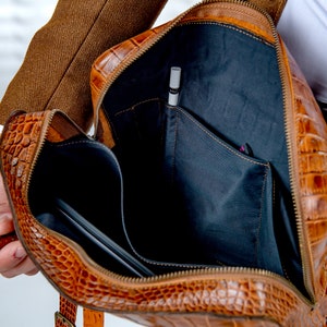 May include: A brown leather briefcase with a crocodile skin effect, open to reveal the interior. The bag has black fabric pockets and a pen. A laptop is visible inside. The zipper and interior details are clearly shown.
