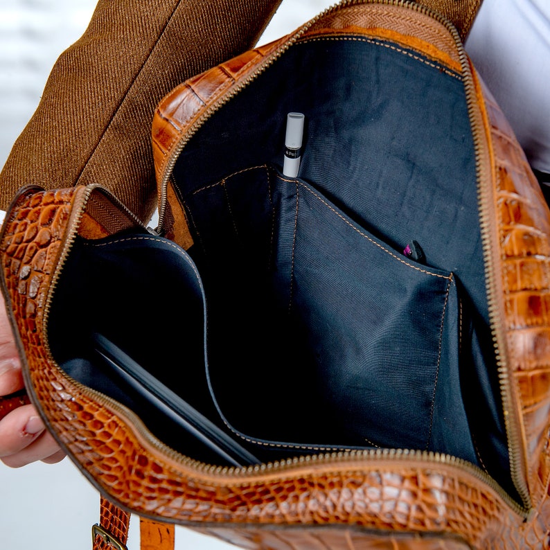May include: A brown leather briefcase with a crocodile skin effect, open to reveal the interior. The bag has black fabric pockets and a pen. A laptop is visible inside. The zipper and interior details are clearly shown.
