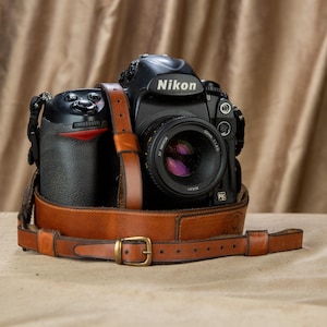 Op de afbeelding: Een zwarte Nikon camera met een 50mm lens en een bruine leren riem. De camera ligt op een oppervlak en de riem is vastgemaakt. Het Nikon logo is zichtbaar.