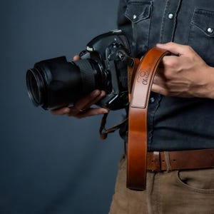 Op de afbeelding: Een bruine leren camerariem met de tekst "Christie Rose" is bevestigd aan een zwarte Nikon camera met een grote lens. De riem wordt vastgehouden door een persoon die een donker spijkeroverhemd en een bruine broek draagt.