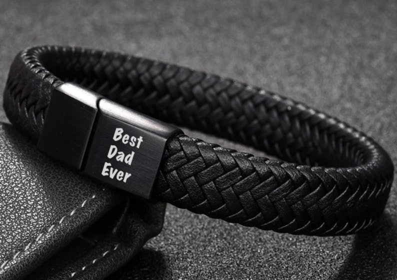 May include: Black braided leather bracelet with a rectangular clasp. The clasp is engraved with the words "Best Dad Ever" in white. The bracelet is a dark, neutral color and appears to be made of woven leather.