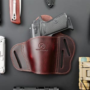 Op de afbeelding: Een donker bordeauxrood leren holster met een zwart pistool. Op het holster staat de merknaam "KOSIBATE" in reliëf. De stiksels zijn zichtbaar en het holster heeft verschillende uitsparingen. Andere gereedschappen zijn zichtbaar op de achtergrond.