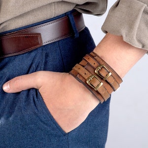 May include: Brown leather cuff bracelet with two straps and brass buckles. The bracelet is worn on the wrist, with a brown leather belt and blue pants in the background. The person is wearing a tan shirt.