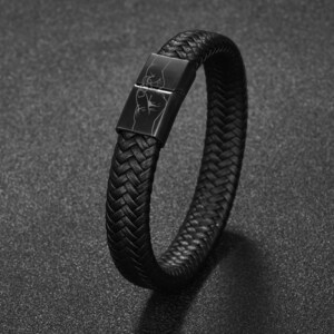 May include: Black braided leather bracelet with a rectangular clasp. The clasp features an etched design of a hand making a fist. The bracelet is a dark, neutral color and appears to be made of woven leather.