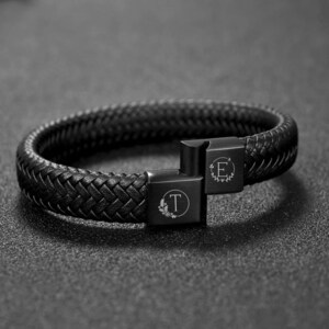 May include: A black leather bracelet with a braided design. The bracelet features a magnetic clasp with the engraved letters "T" and "E" surrounded by floral accents. The bracelet is shown on a dark surface.