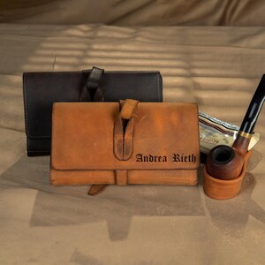 May include: Two leather tobacco pouches, one brown and one tan, with a clasp closure. The tan pouch has the name "Andrea Rieth" printed on it. A wooden pipe and a pipe stand are also visible.