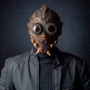 May include: A brown and tan leather mask with brass accents and goggles. The mask has a steampunk aesthetic, with rivets and a stitched design. The person is wearing a black jacket and a black turtleneck.