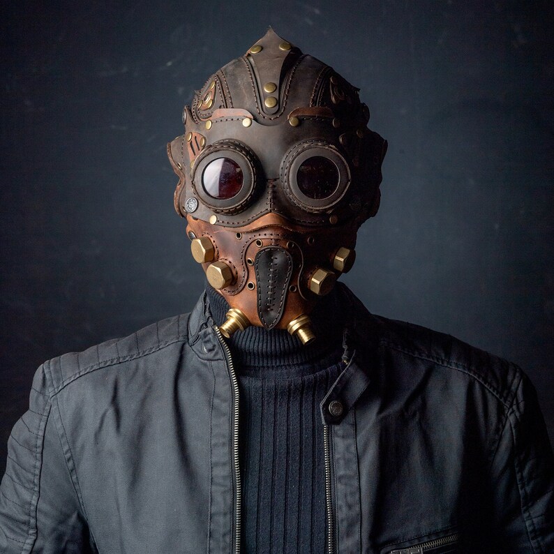 May include: A brown and tan leather mask with brass accents and goggles. The mask has a steampunk aesthetic, with rivets and a stitched design. The person is wearing a black jacket and a black turtleneck.