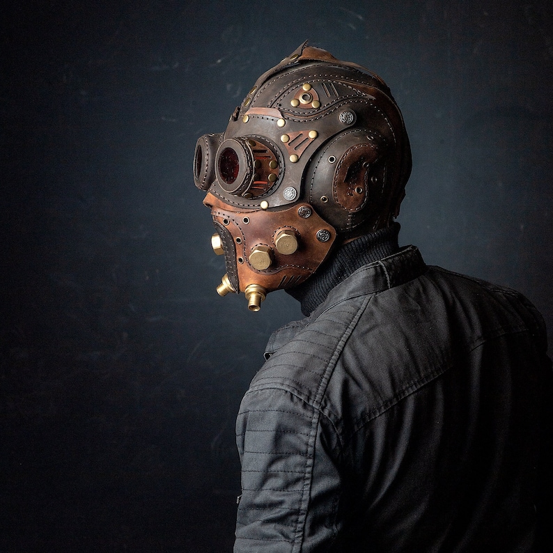 May include: A brown and tan leather steampunk mask with brass accents and red-tinted goggles. The mask features intricate stitching and rivet details, with a design that covers the entire head and face.