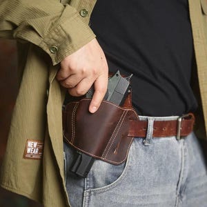 May include: A brown leather holster with a gun inside, worn on the waist with a brown belt and blue jeans. The holster is stitched with white thread. The person is wearing a green shirt and a black shirt.