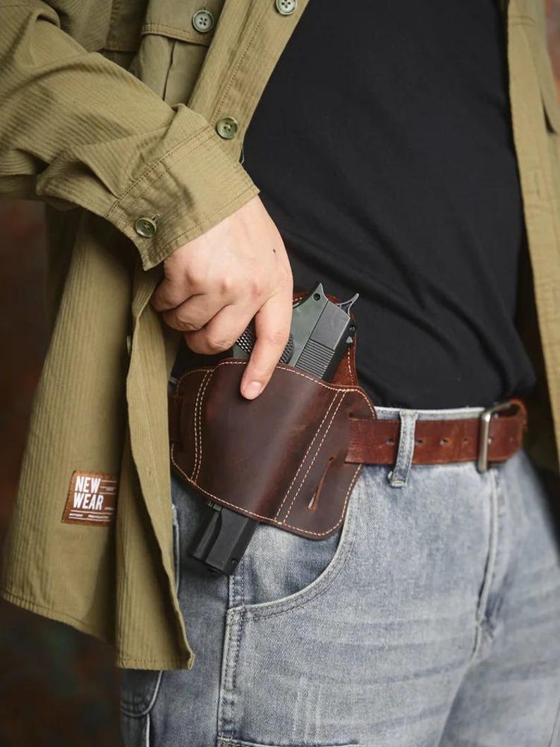 May include: A brown leather holster with a gun inside, worn on the waist with a brown belt and blue jeans. The holster is stitched with white thread. The person is wearing a green shirt and a black shirt.