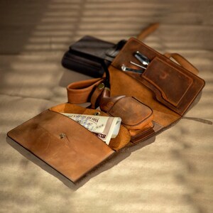 May include: A collection of brown leather accessories, including a tobacco pipe case, a pouch, and a wallet. The wallet is open, revealing a stack of paper currency. The accessories are arranged on a light brown surface.