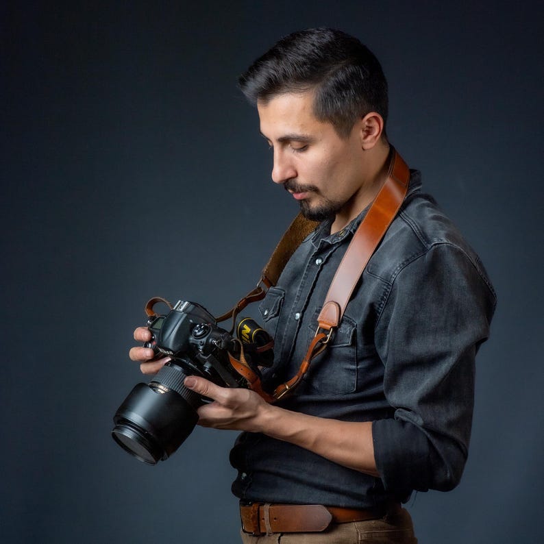 Op de afbeelding: Een zwarte DSLR-camera met lens wordt vastgehouden. De camera heeft een bruine leren riem. De persoon draagt een donkergrijs overhemd met knopen en een bruine broek. De achtergrond is donker.