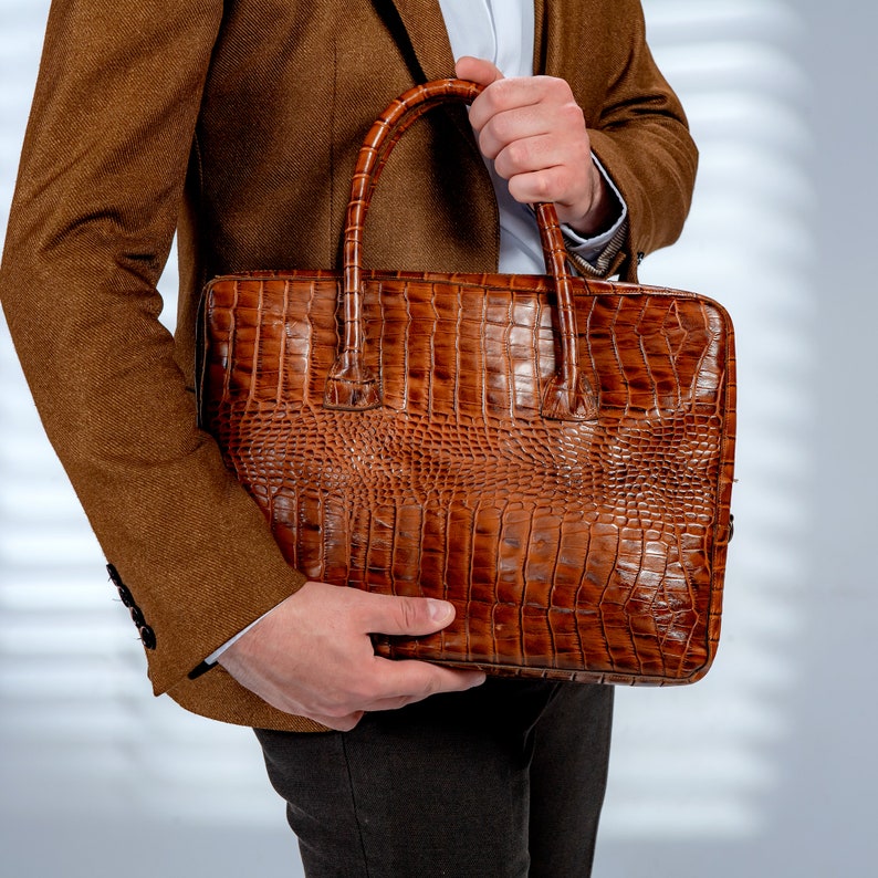 May include: A brown leather briefcase with a crocodile skin effect. The briefcase has a rectangular shape with a handle on top. The colour is a rich, warm brown. The briefcase is held by a person wearing a brown blazer.