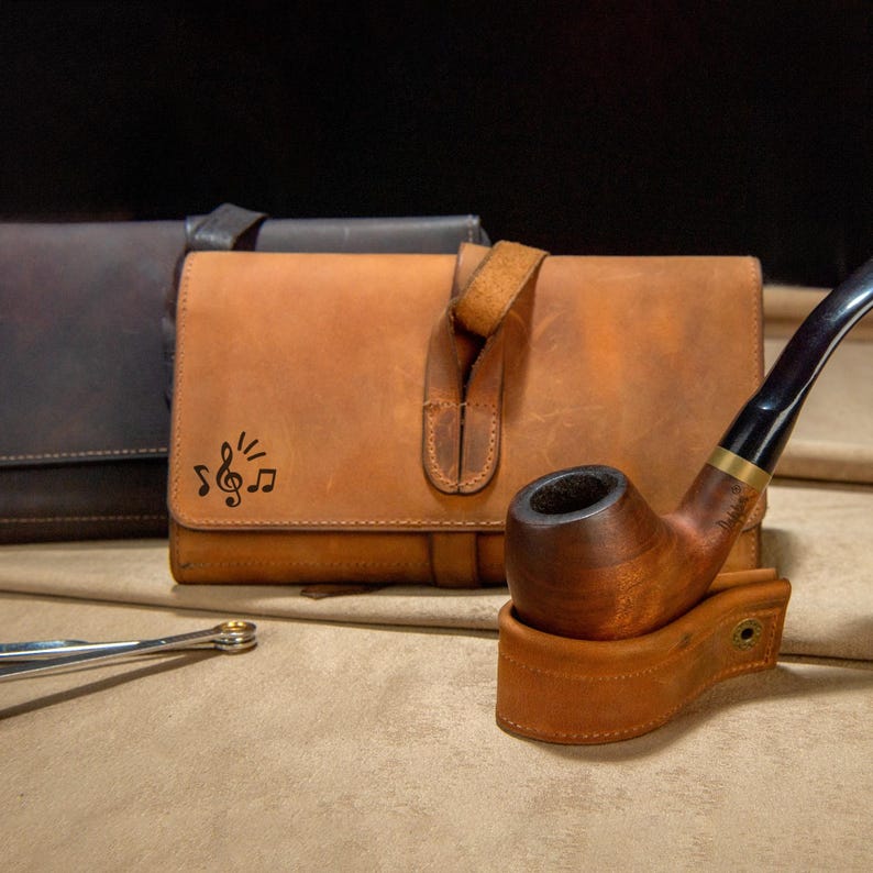 May include: A wooden tobacco pipe with a curved stem rests on a leather stand. A brown leather pouch with a musical note design is in the background. A silver tool is in the foreground. The items are on a tan surface.