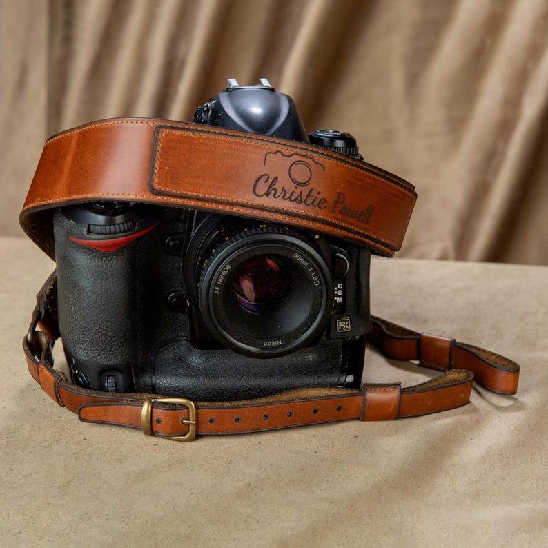 Op de afbeelding: Een zwarte DSLR-camera met een bruine leren riem. De riem is voorzien van de naam "Christie Powell" en een camera-icoon. De cameralens is zwart met zilveren accenten. De riem heeft een messing gesp.