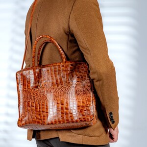 May include: A brown, textured briefcase with a crocodile skin pattern. The bag has a top handle and a shoulder strap. The briefcase is carried over the shoulder of a person wearing a brown blazer.