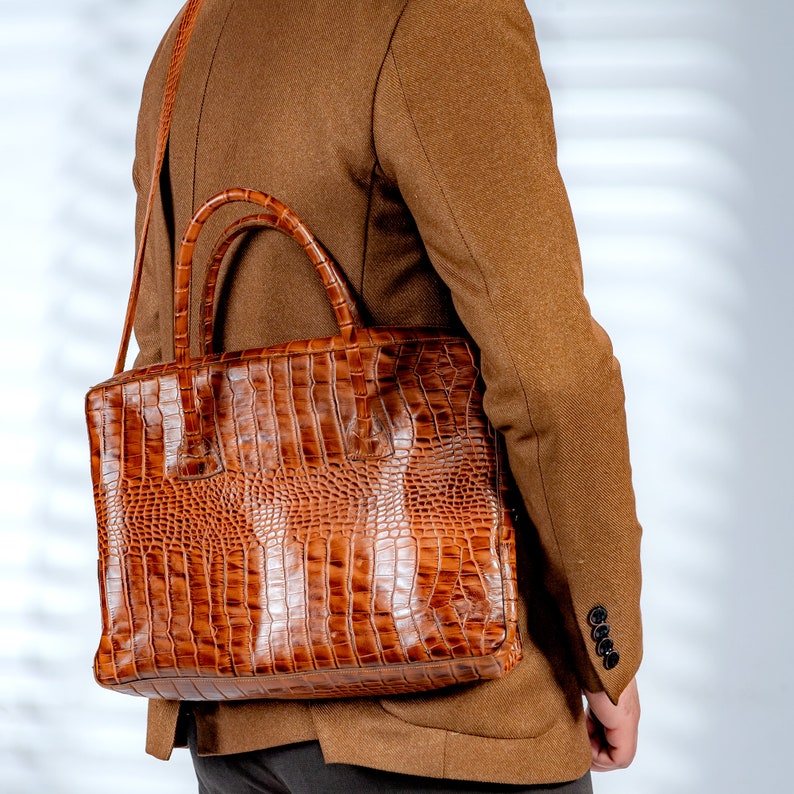 May include: A brown, textured briefcase with a crocodile skin pattern. The bag has a top handle and a shoulder strap. The briefcase is carried over the shoulder of a person wearing a brown blazer.