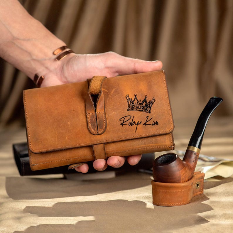 May include: A brown leather tobacco pipe case and a wooden pipe with a black stem. The case has a crown graphic and the name "Robyn Kim" printed on it. The pipe is in a matching leather holder.