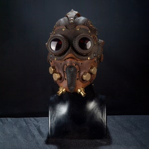 May include: A brown and tan leather steampunk mask with large, round goggles and brass accents. The mask features intricate stitching, rivets, and a beak-like nose piece, creating a unique and detailed design. The goggles have red lenses.