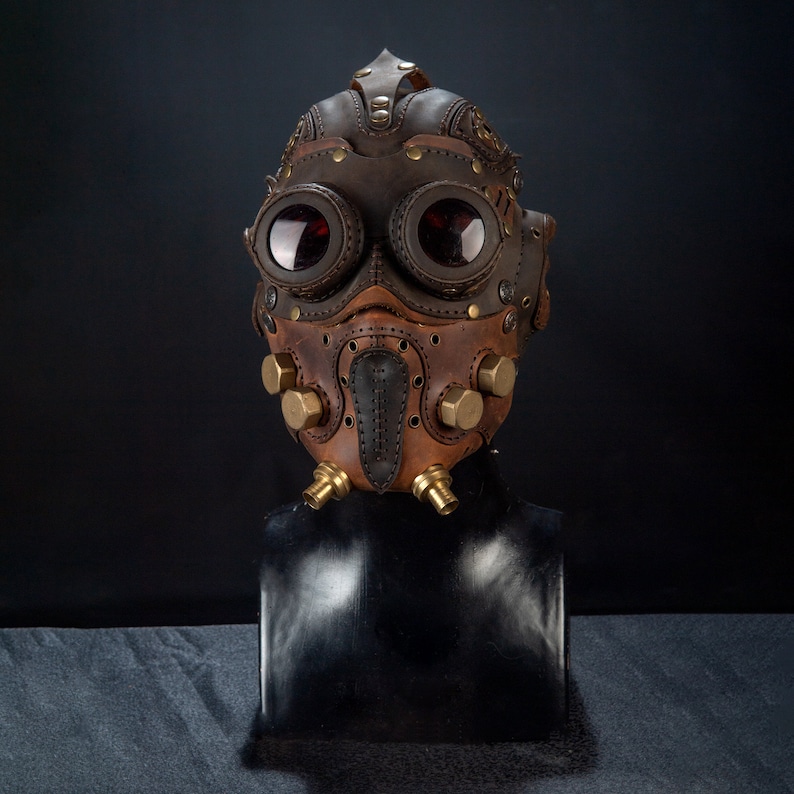 May include: A brown and tan leather steampunk mask with large, round goggles and brass accents. The mask features intricate stitching, rivets, and a beak-like nose piece, creating a unique and detailed design. The goggles have red lenses.