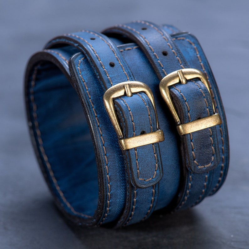 May include: A blue leather cuff bracelet with two straps and gold-colored buckles. The bracelet is stitched with a contrasting thread and has a distressed finish. The cuff is designed to wrap around the wrist.