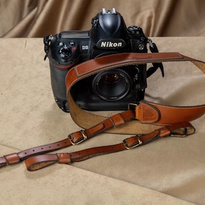 Op de afbeelding: Een zwarte Nikon D3 digitale SLR-camera met een bruinleren camerariem. De riem heeft messing gespen en ligt op een bruine ondergrond. De cameralens is zichtbaar.